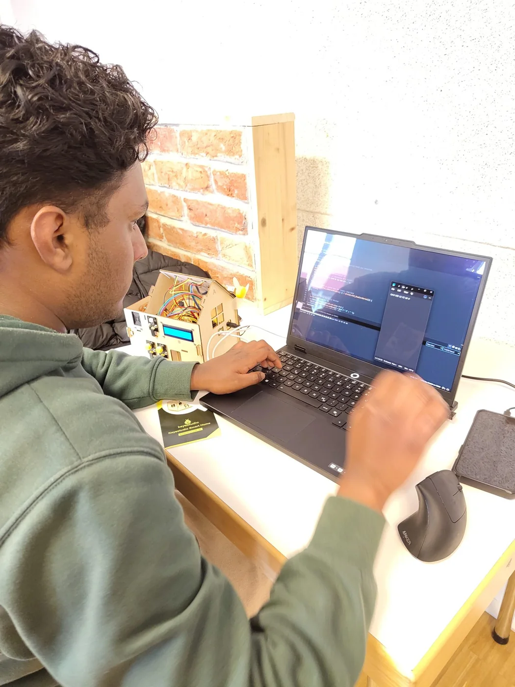 A participant writing code on a laptop next to a small model building rigged with electronics